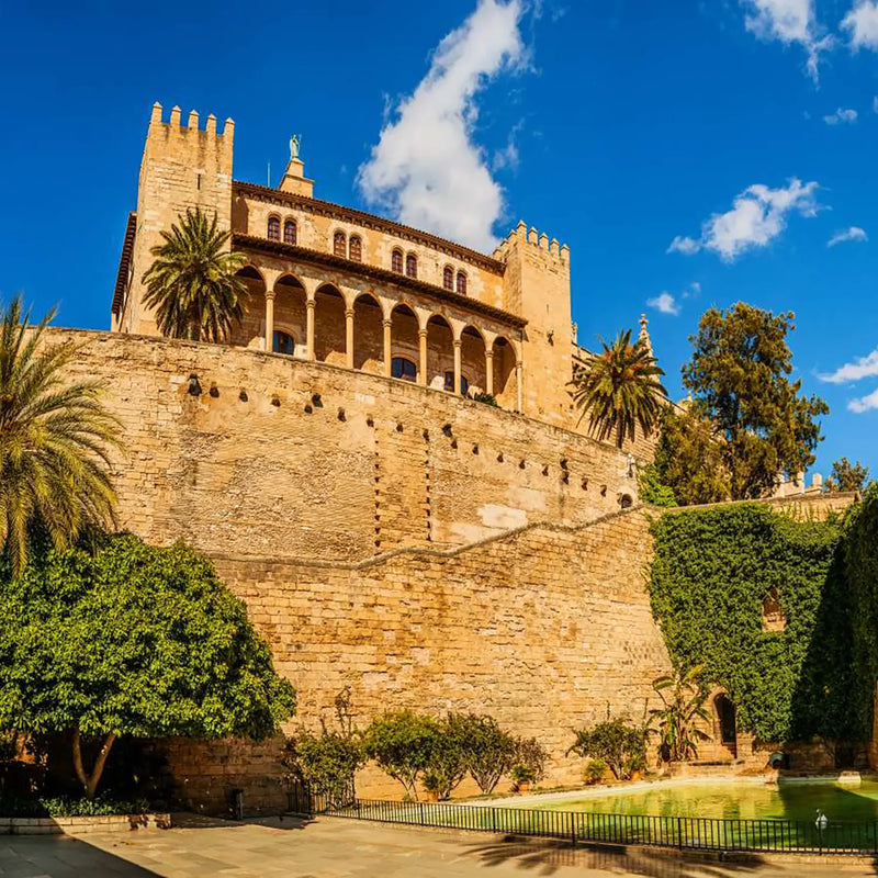 Vista del Palacio Real de la Almudaina en Palma de Mallorca, con murallas de piedra, palmeras y cielo azul con nubes.
