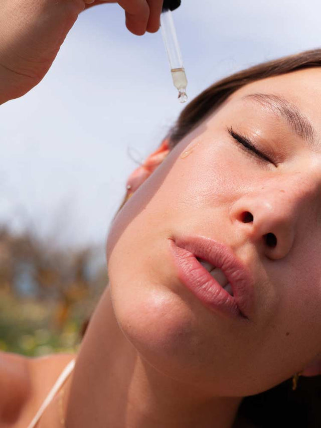 Mano aplicando gotas de serum en un frasco marrón y mujer con ojos cerrados recibiendo el producto en su mejilla.