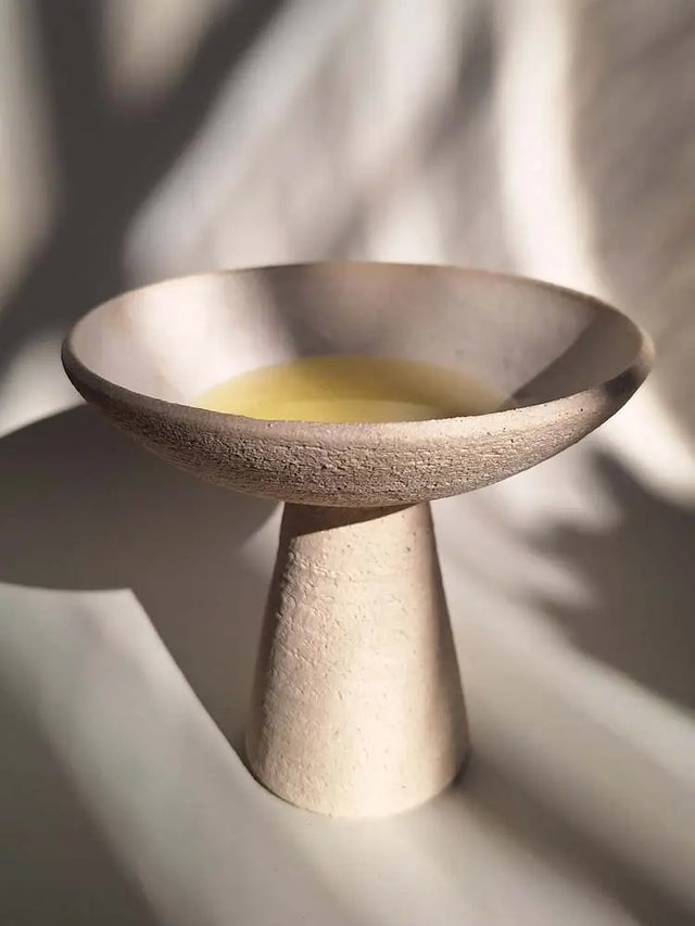 Cuenco decorativo de cerámica beige con pedestal para ofrendas o aceites. Diseño moderno y minimalista bajo luz natural.