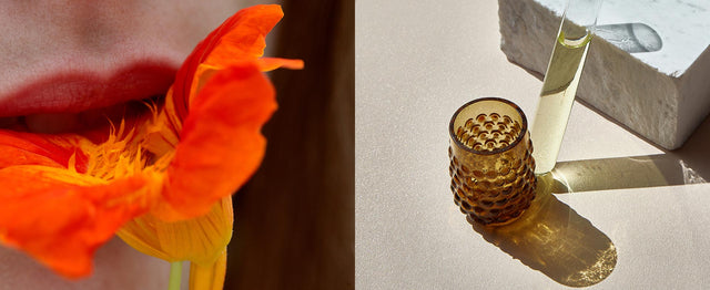 Labios sosteniendo una flor naranja vibrante, junto a una botella y un vaso de vidrio ámbar sobre una superficie iluminada.