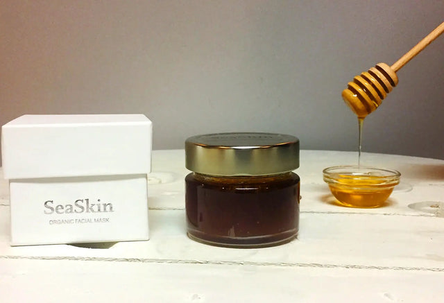 Un frasco pequeño de vidrio oscuro con tapa dorada lleno de miel, junto a una caja blanca de la marca SeaSkin. Un cucharón de miel de madera gotea el líquido en un cuenco pequeño de vidrio. Los productos están sobre una superficie de madera clara.