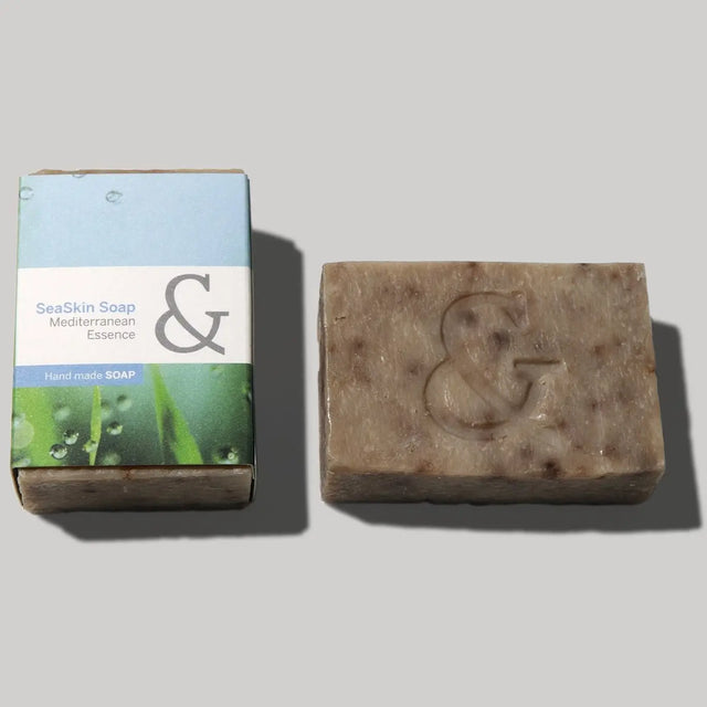 Pastilla de jabón artesanal de color marrón claro, con el relieve de un signo de 'ampersand' (&) en el centro, junto a su caja de cartón de color azul y verde con el texto 'SeaSkin Soap Mediterranean Essence Handmade Soap'.