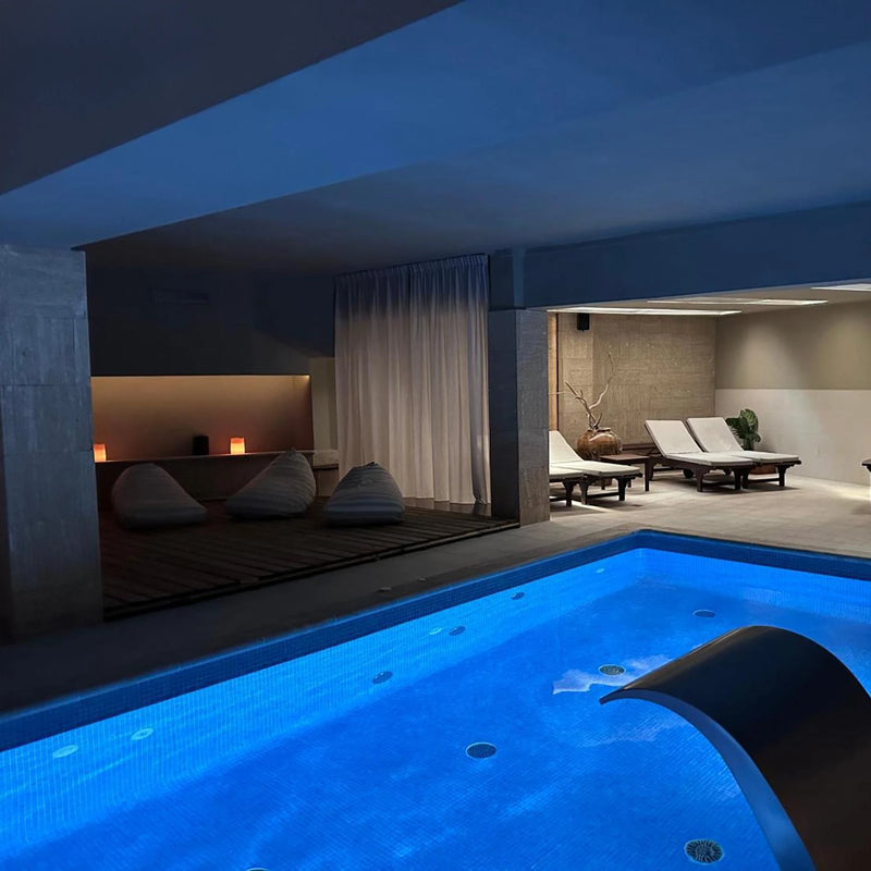 Piscina interior iluminada en tonos azules, con cascada metálica, tumbonas de relax y zona de descanso con pufs y luz tenue en un spa elegante.