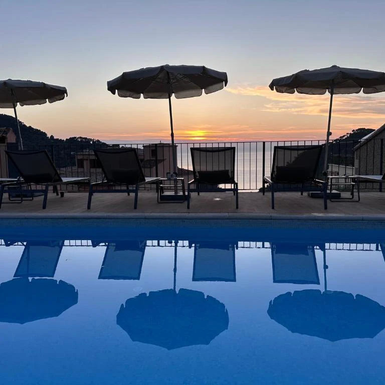 Piscina exterior con tumbonas y sombrillas, reflejando un atardecer sobre el mar Mediterráneo en un entorno tranquilo y montañoso.