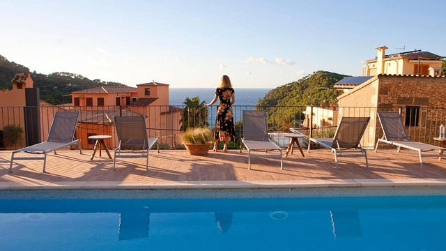 Mujer de pie junto a una barandilla en una terraza con tumbonas, contemplando el mar al atardecer desde un hotel en la sierra de Mallorca.