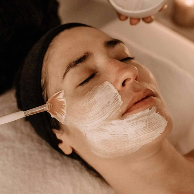 Aplicación de una mascarilla facial blanca con una brocha durante un tratamiento de relajación (relax) en un spa.