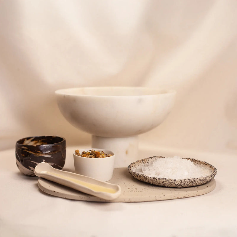 Set de spa con cuencos de cerámica que contienen sal marina, flores secas y productos naturales, sobre fondo beige suave.