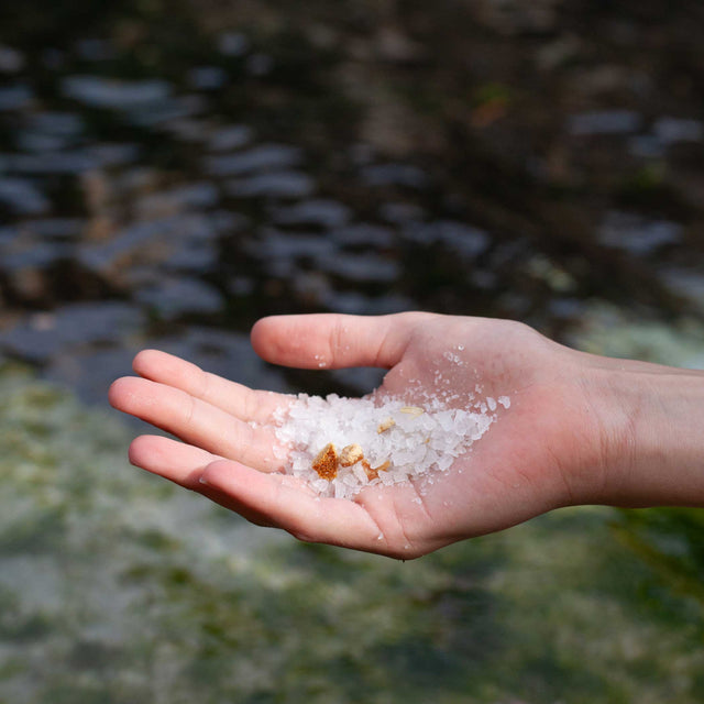 Mano sosteniendo sal marina frente a un agua cristalina, simbolizando pureza y origen natural.