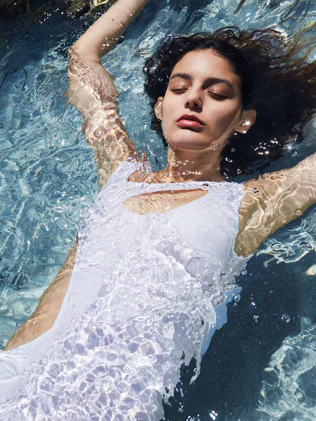 Mujer flotando en el agua con los ojos cerrados, lleva traje de baño blanco y se ve relajada bajo el sol.