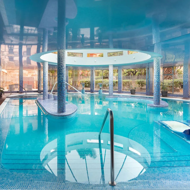 Piscina interior de diseño elegante con columnas recubiertas de mosaicos azules y luz natural que entra a través de amplias ventanas panorámicas, rodeada de vegetación exterior.