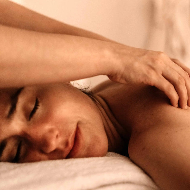 Mujer relajada y sonriente con los ojos cerrados disfrutando de un tratamiento corporal o masaje, con una mano sobre el hombro.