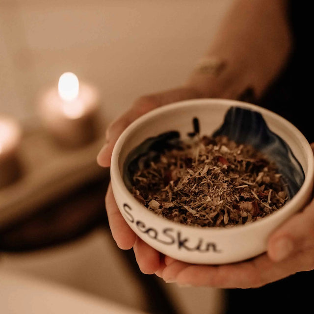 Primer plano de manos sosteniendo un cuenco de cerámica blanca SeaSkin con mezcla de hierbas secas naturales para tratamiento facial o corporal de spa. ALQUIMISTISCHE KÖRPERMASSAGE | Can Marqués Palma