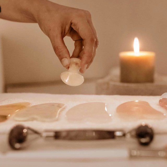 Primer plano de una mano levantando una ventosa de masaje facial de silicona o vidrio, con varias herramientas de Gua Sha de cuarzo rosa y jade, y un rodillo facial, sobre una toalla blanca. Una vela encendida ilumina la escena.