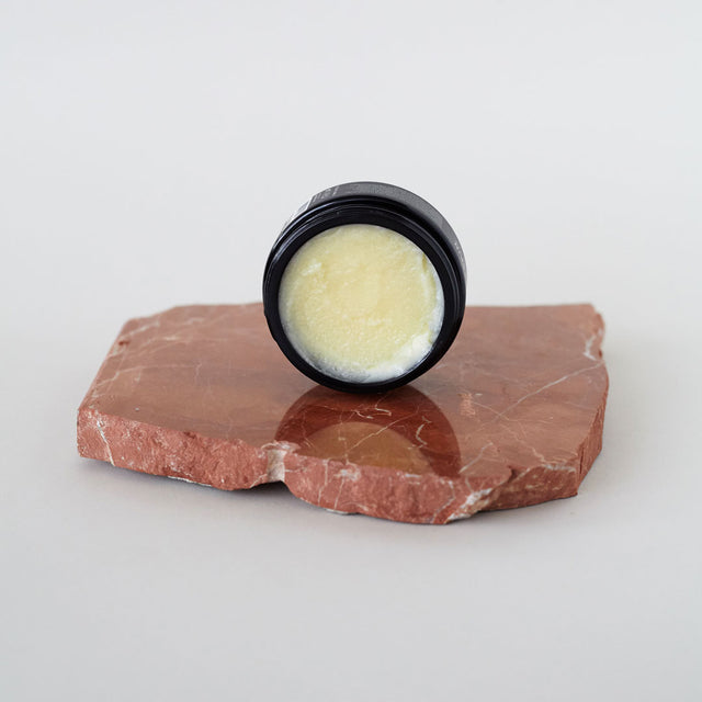 Envase de bálsamo facial desmaquillante de Beseaskin, 100% orgánico a base de manteca de mango y cacao, sobre piedra en una mesa blanca.
