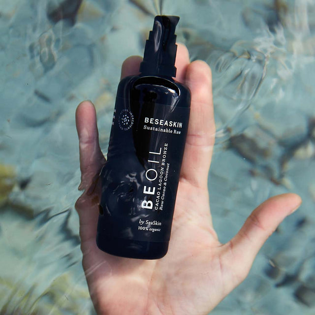 Envase de aceite corporal bronceador de Beseaskin, 100% orgánico a base de Raw Cacao y Coco en una mano dentro del mar.