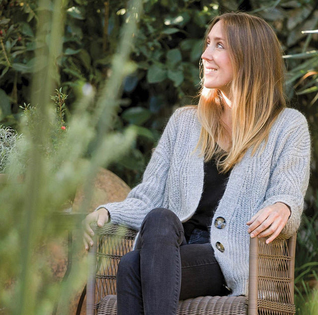 Imagen de Paloma Ruiz sentada en una terraza con plantas de fondo, sonriente, mirando al horizonte.