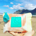 Neceser con estampado de lengua mallorquina de color blanco, naranja y amarillo y logo de SeaSkin Life de color turquesa en el centro, con paisaje de playa de fondo.