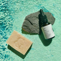 Envase de Elixir Ío, aceite facial hidratante de Smell The Planet, junto a Jabón Vegetal Artesanal regenerador de SeaSkin sobre superficie rocosa verde turquesa